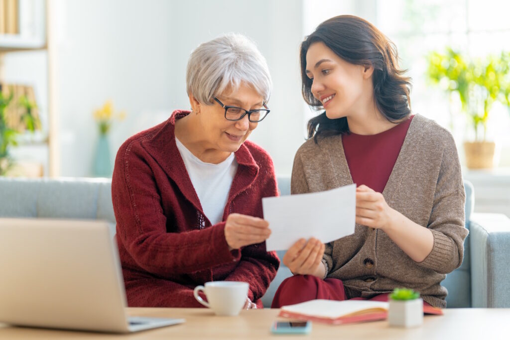 Two women, sitting on the sofa with a paper receipt, are calculating expenses, managing the family budget.