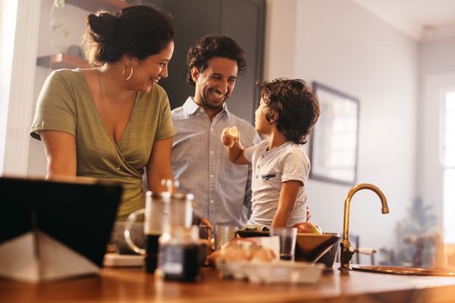 Family time in the kitchen. Happy young boy eating a banana while sitting on a kitchen counter. Mom and dad smiling happily while spending quality time with their son at home.