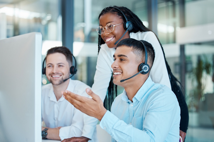 Three smiling people wearing headsets and looking at a computer monitor.