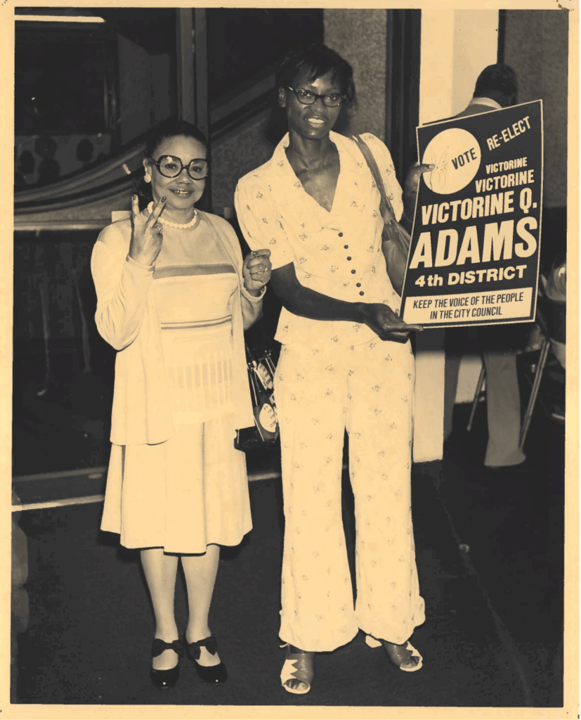 A black and white photo of two people holding a Re-elect Victorine Adams sign