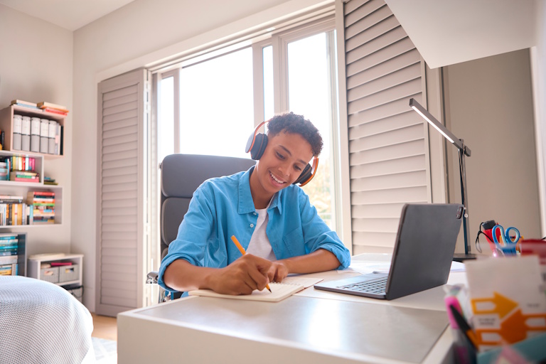 Teenage Boy Sitting At Desk In Bedroom At Home Doing Homework Streaming Music To Headphones
